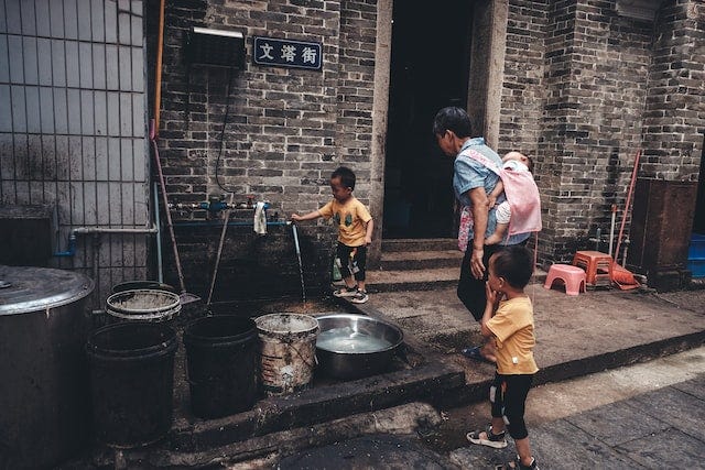 Foto de criança chinesa abrindo uma torneira de água do lado de fora de uma casa chinesa de tijolos cinzas. Com a criança há uma senhora, carregando um bebê no corpo e de mão dada com outro menino. Foto de criança chinesa abrindo uma torneira de água do lado de fora de uma casa chinesa de tijolos cinzas. Com a criança há uma senhora, carregando um bebê no corpo e de mão dada com outro menino.