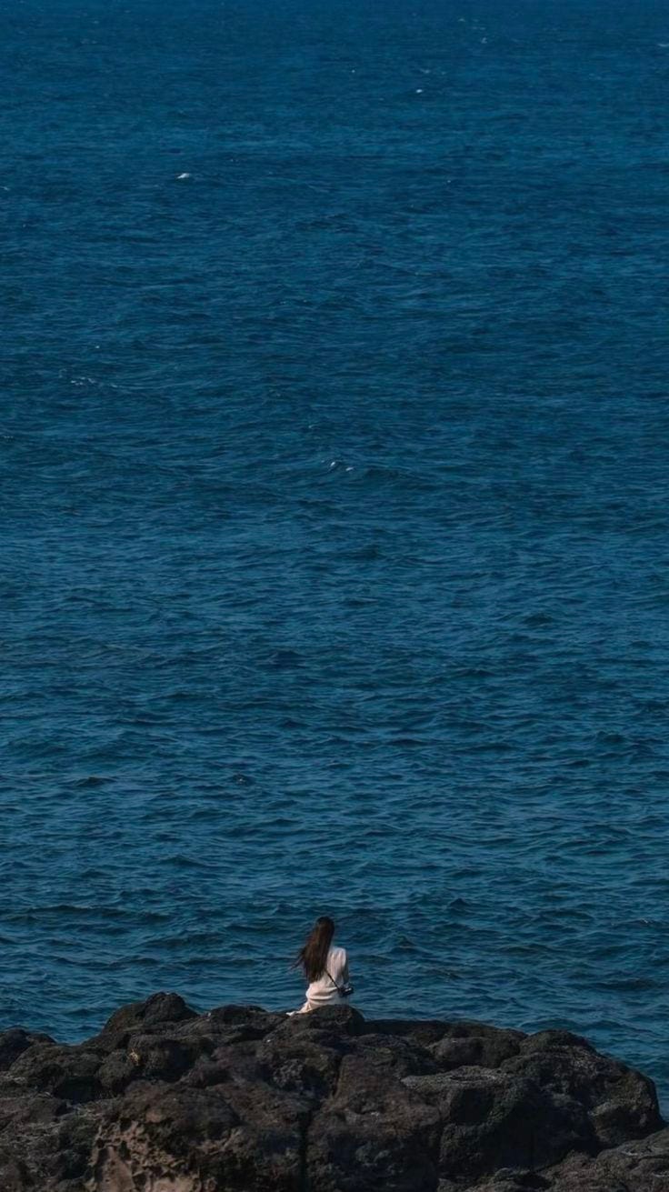 This may contain: a person sitting on rocks near the ocean with a kite flying in the sky above them