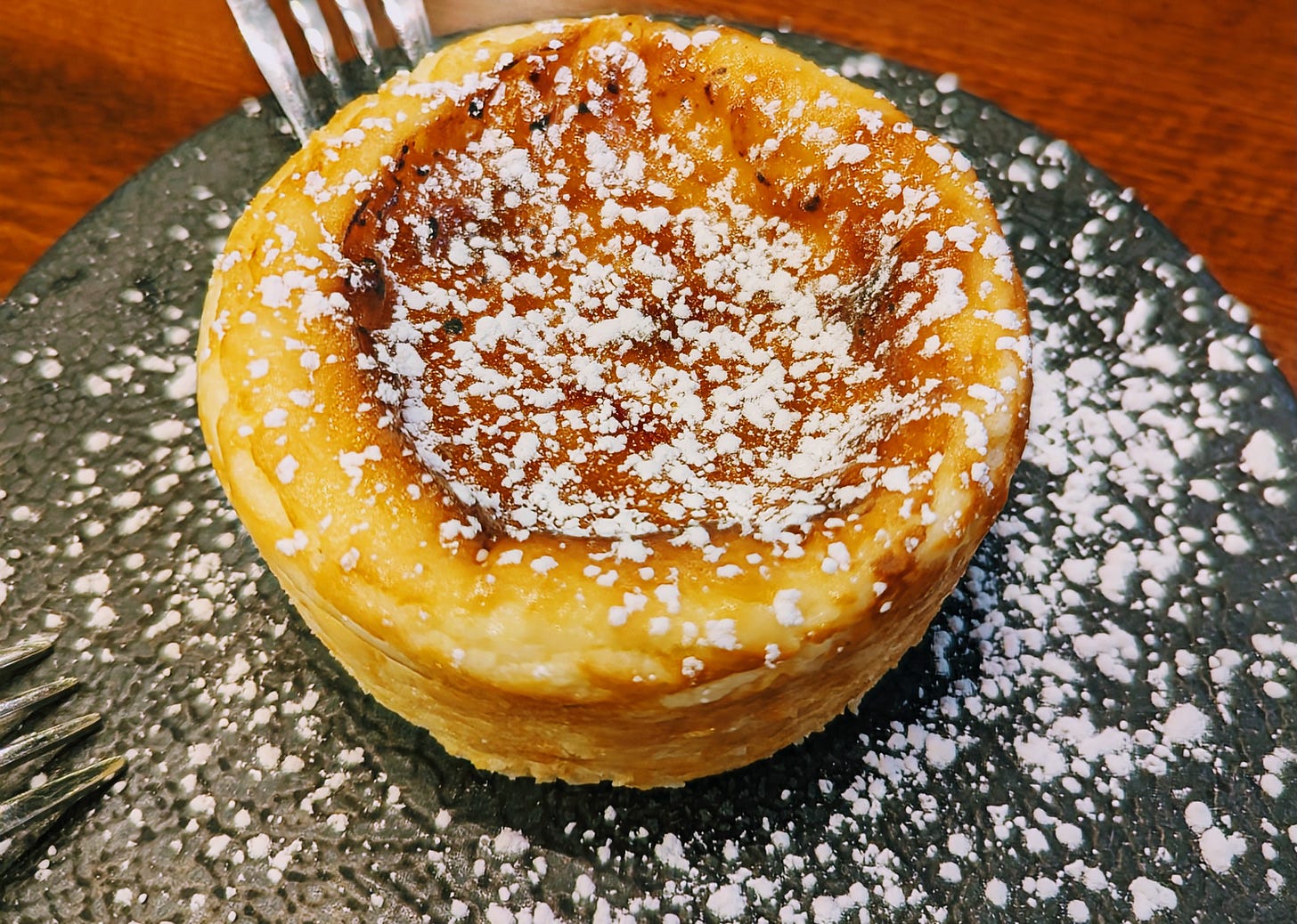 Creme brûlée cheesecake topped with powdered sugar on a black plate.