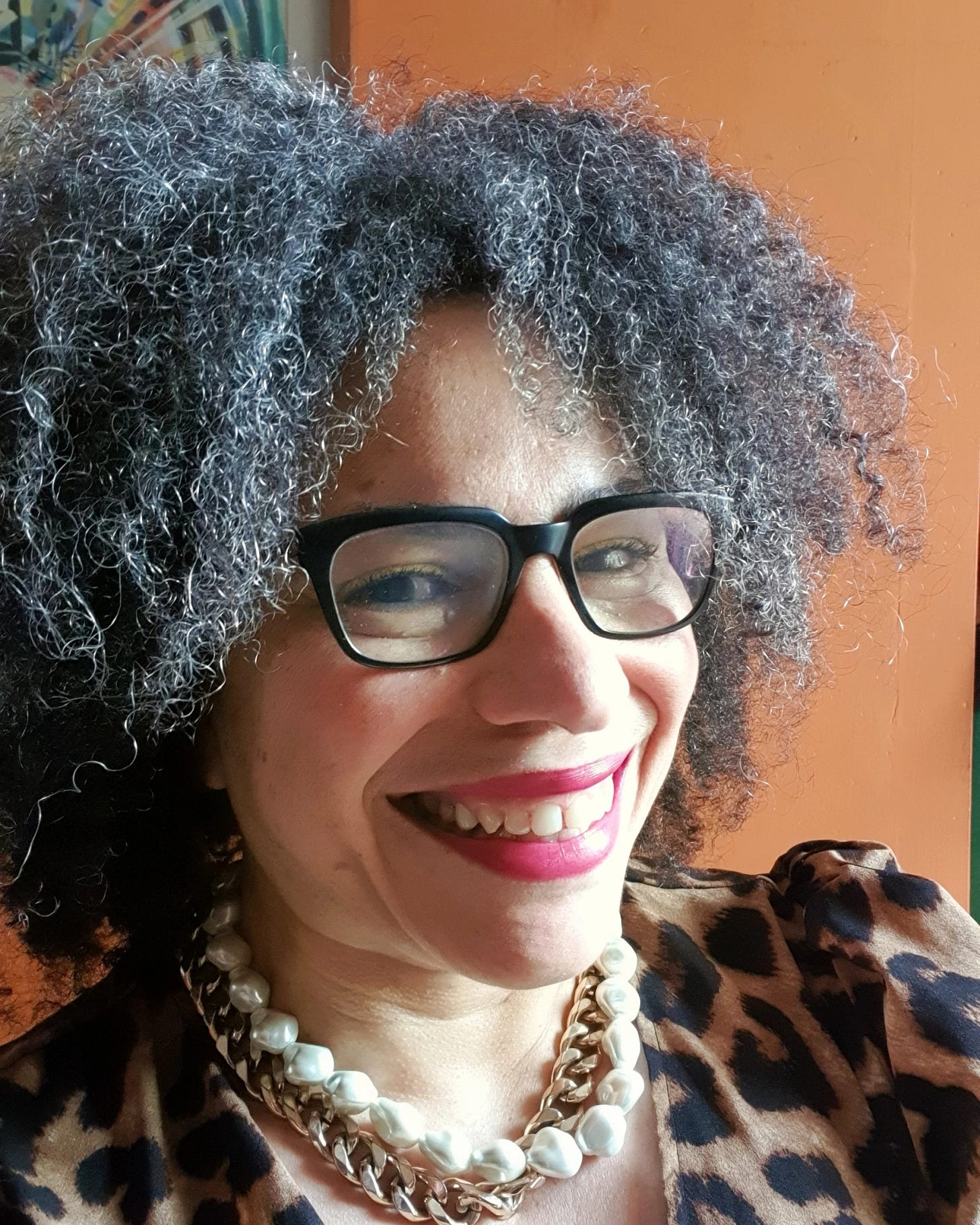 Picture of a middle-aged woman looking at the camera and smiling. She is wearing glasses, bright red lipstick, a leopard print dress and a chunky gold chain and fake pearl necklace. She looks vibrant and happy.