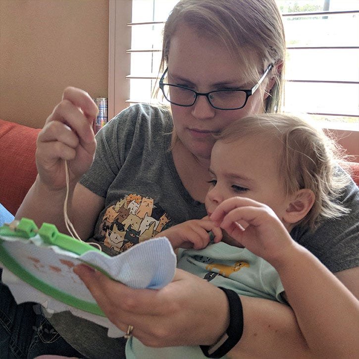 An older photo of Kate from Two Little Kits with her youngest daughter (when she was a toddler) in her lap, watching Kate stitch a piece. An older photo of Kate from Two Little Kits with her youngest daughter (when she was a toddler) in her lap, watching Kate stitch a piece.