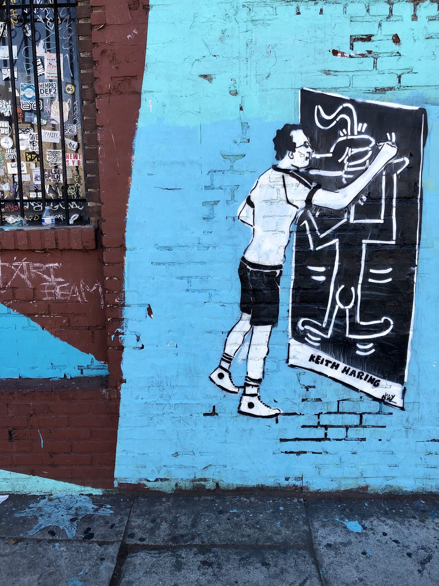 Street art on a blue brick wall shows an artist painting a Keith Haring-style figure. A nearby window features numerous stickers. The figure is supposed to be Keith Haring doing one of his own street arts. It's a classic Keith figure with a black background and white outline, and his signature on the bottom. Street art on a blue brick wall shows an artist painting a Keith Haring-style figure. A nearby window features numerous stickers. The figure is supposed to be Keith Haring doing one of his own street arts. It's a classic Keith figure with a black background and white outline, and his signature on the bottom.