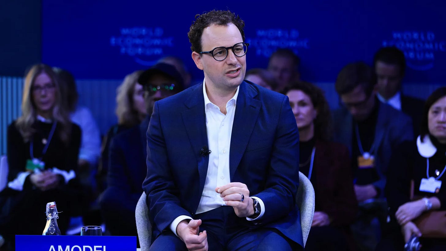 Man seated in a suit and white shirt, no tie, with an audience seated behind him. 