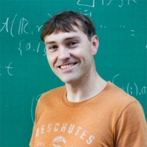 20260715_eddienijholt Homem de cabelos castanhos sorridente, usando camiseta laranja e calça bege, em frente a uma lousa com cálculos matemáticos