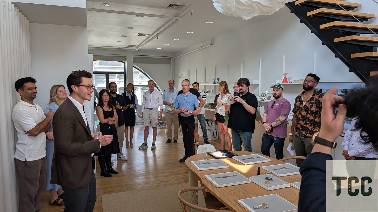 Teddy Baldassarre, dressed in a dark blazer, speaks to a semicircle of guests at the NOMOS Lounge. Attendees stand with drinks in hand, listening attentively beneath soft lighting and floating lighting features.