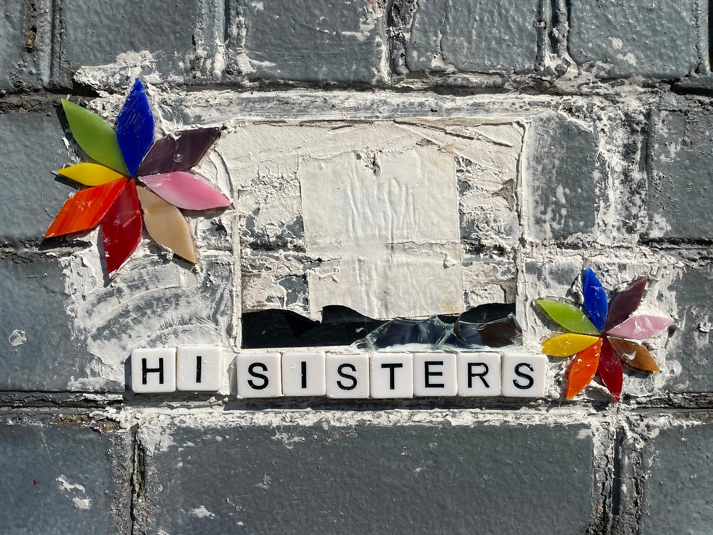 grout on gray brick wall, into which are pressed two rainbow flowers made of tile and letter tiles that say, Hi Sisters.