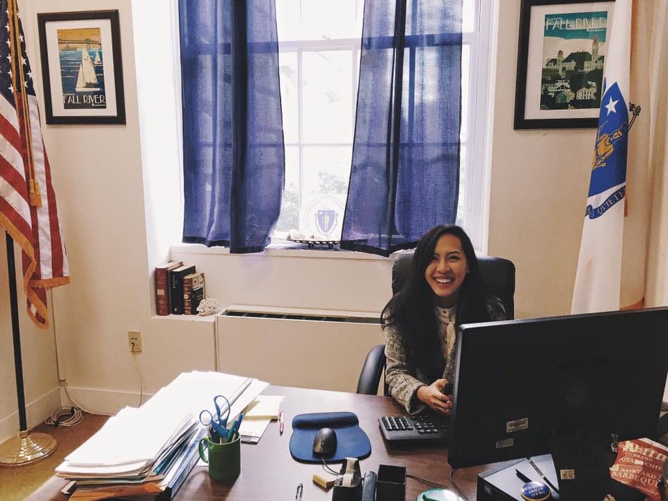 A young woman sits smiling at a large wooden desk in an office, surrounded by state flags and paperwork. Sunlight filters through navy curtains behind her, highlighting official-looking decor and framed posters on the wall.