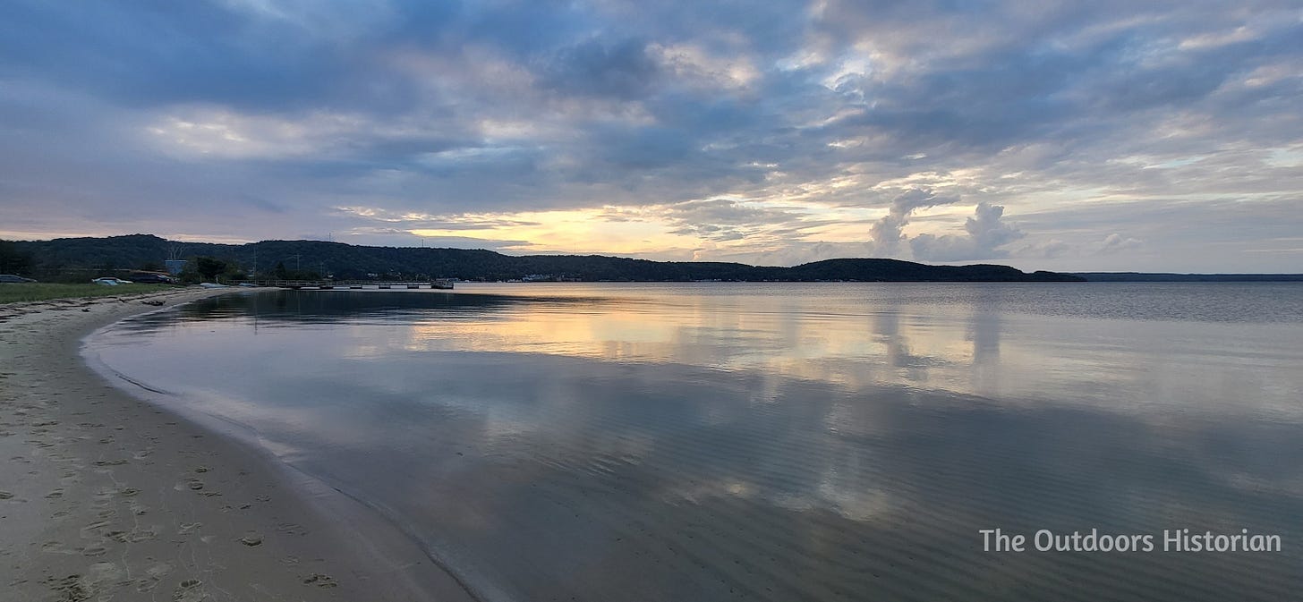 A golden sun illuminates the clouds and reflects on a calm Lake Superior with the hill over the city of Munising in the distance.