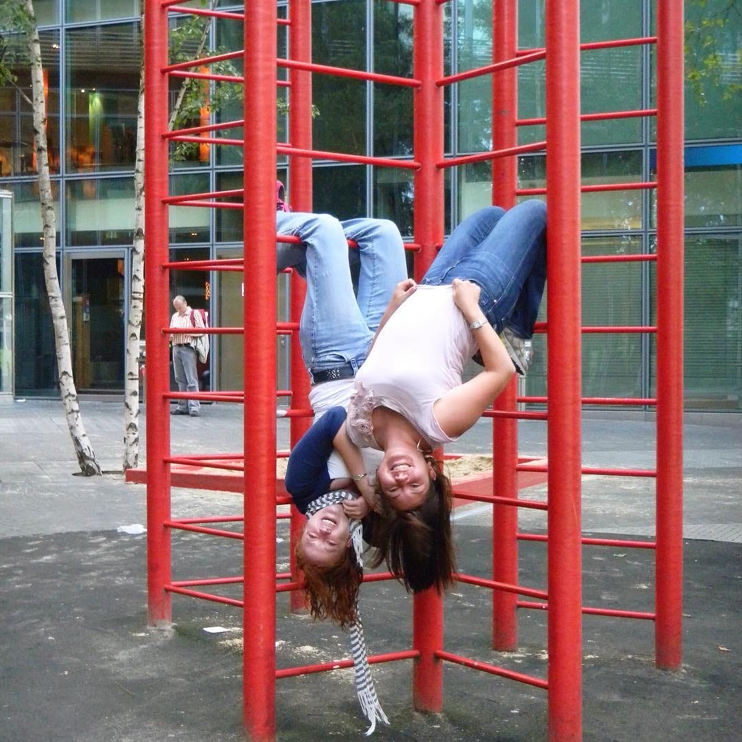 Photo of two young women hanging upside down from a red ladder-like structure outdoors. Both are wearing jeans and grinning, their hair dangling beneath them.