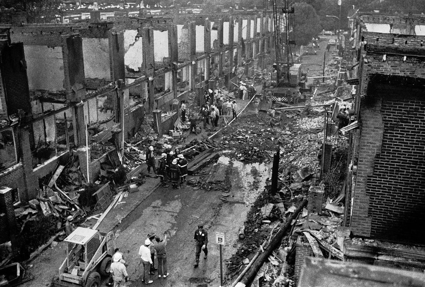 Black and white aerial photo of the street that the MOVE Bombing took place in | Philadelphia, Pennsylvania USA. There is debris everywhere and a row of brick buildings have the insides of them completely blown out. People stand on the street, walking and pointing. Black and white aerial photo of the street that the MOVE Bombing took place in | Philadelphia, Pennsylvania USA. There is debris everywhere and a row of brick buildings have the insides of them completely blown out. People stand on the street, walking and pointing.