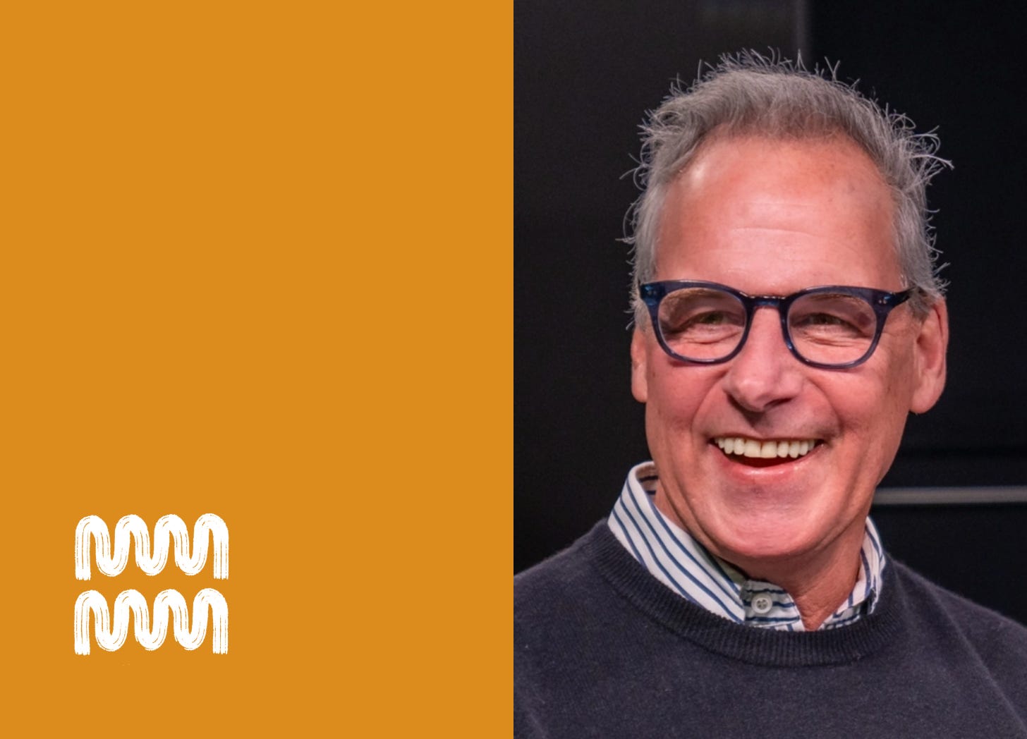 Mark has gray hair and wears blue framed glasses. He wears a blue striped shirt and a gray sweater. He is smiling mid-sentence. Mark has gray hair and wears blue framed glasses. He wears a blue striped shirt and a gray sweater. He is smiling mid-sentence.