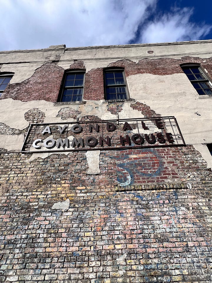 a brick building on a corner with blue skies behind it and a metal sign on a brick wall that says Avondale Common House