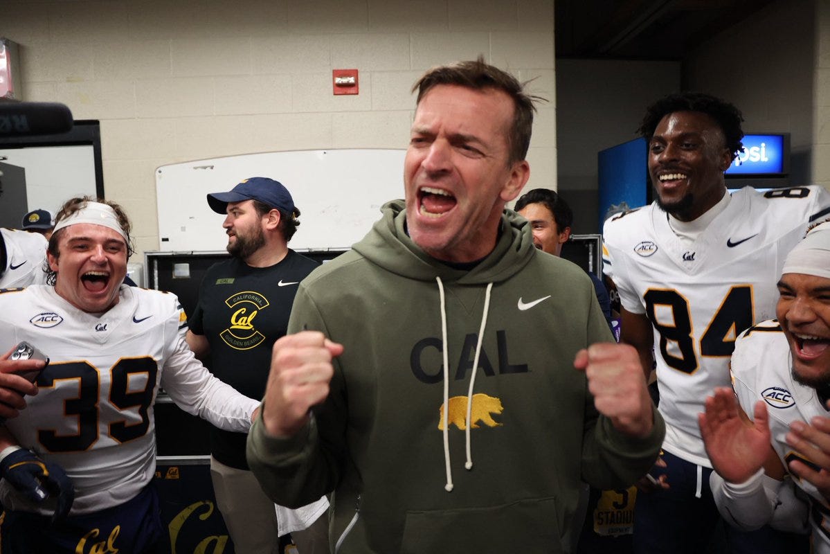 Several young men in white and gold American football uniforms with numbers 39, 84, and 91 stand smiling and cheering in a locker room. A man in a green hoodie with Cal Bears logo raises both arms excitedly. Another man in a cap claps nearby. Background includes white walls, a whiteboard, and overhead lights.