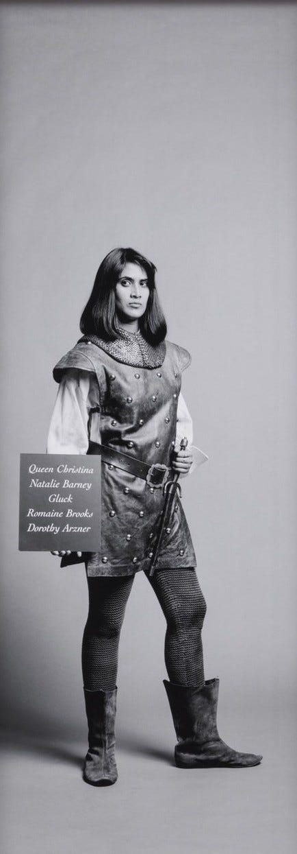 1) A woman stands confidently in full medieval armor, featuring polished metal plates and chainmail. The grayscale image has a serious and heroic tone. The person is white with short hair, and she is standing with her armored hands on her hips. There is a sword attached to her right hip. She does not have a helmet, but she does have metal boots. 2) A brown woman in medieval armor with a confident pose, standing against a plain backdrop. The black-and-white image conveys a serious, historical tone. She has shoulder-length dark hair, and she is wearing a white long-sleeve undershirt, a vest, a belt with large buckle, dark pants, and dark boots. On her right arm, there is a sign that includes the names: 'Queen Christina, Natalie Barney, Gluck, Romaine Brooks, and Dorothy Arzner', which are famous historical lesbians and/or artists. She is also holding a dagger in front of her. Her vest is textured and her pants are chainmail.