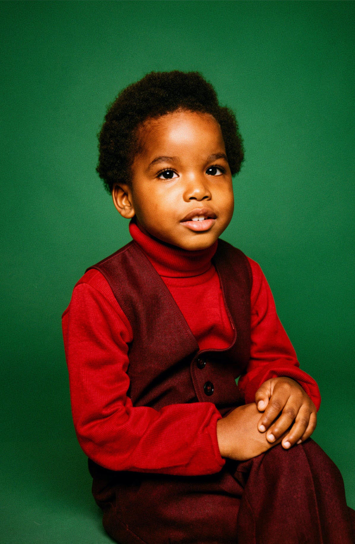 A young Black boy wearing a red shirt under a maroon wool vest and pants, against a deep green background.
