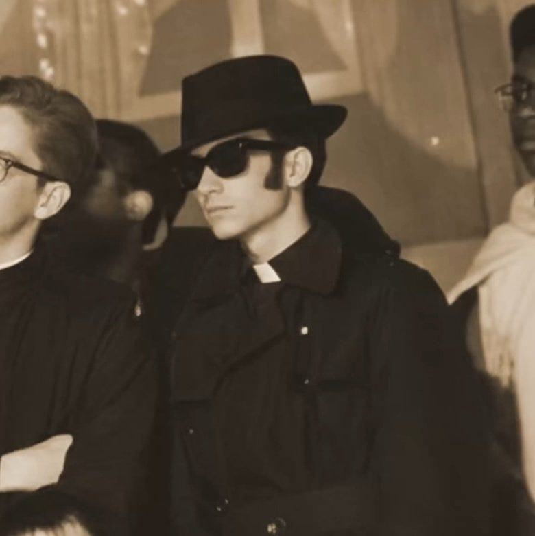 Sepia-toned photograph of a man wearing a black fedora hat, dark sunglasses, and a black clerical collar with cassock, standing in a group with other individuals in similar dark attire, set against draped curtains and soft lighting.
