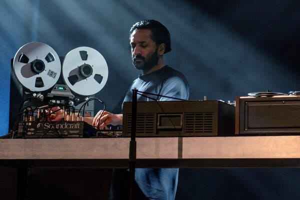 A bearded man stands over a table containing audio equipment, his gaze cast downward at his hands touching knobs, as a reel-to-reel machine is visible behind him. A bearded man stands over a table containing audio equipment, his gaze cast downward at his hands touching knobs, as a reel-to-reel machine is visible behind him.