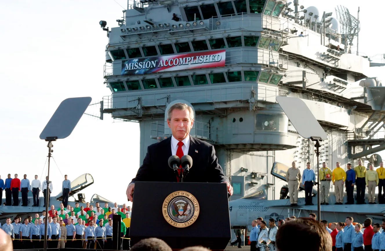 George W. Bush speaks from a podium with a "Mission Accomplished" banner on the USS Abraham Lincoln.