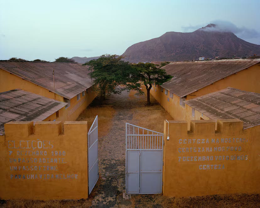 Ingresso del campo di concentramento di Tarrafal: un cancello in ferro incorniciato da muri perimetrali gialli delle caserme, con un viale centrale di terra battuta che conduce verso l’interno. Ai lati del viale si trovano due alberi e, sullo sfondo, si intravede una montagna sotto un cielo chiaro.