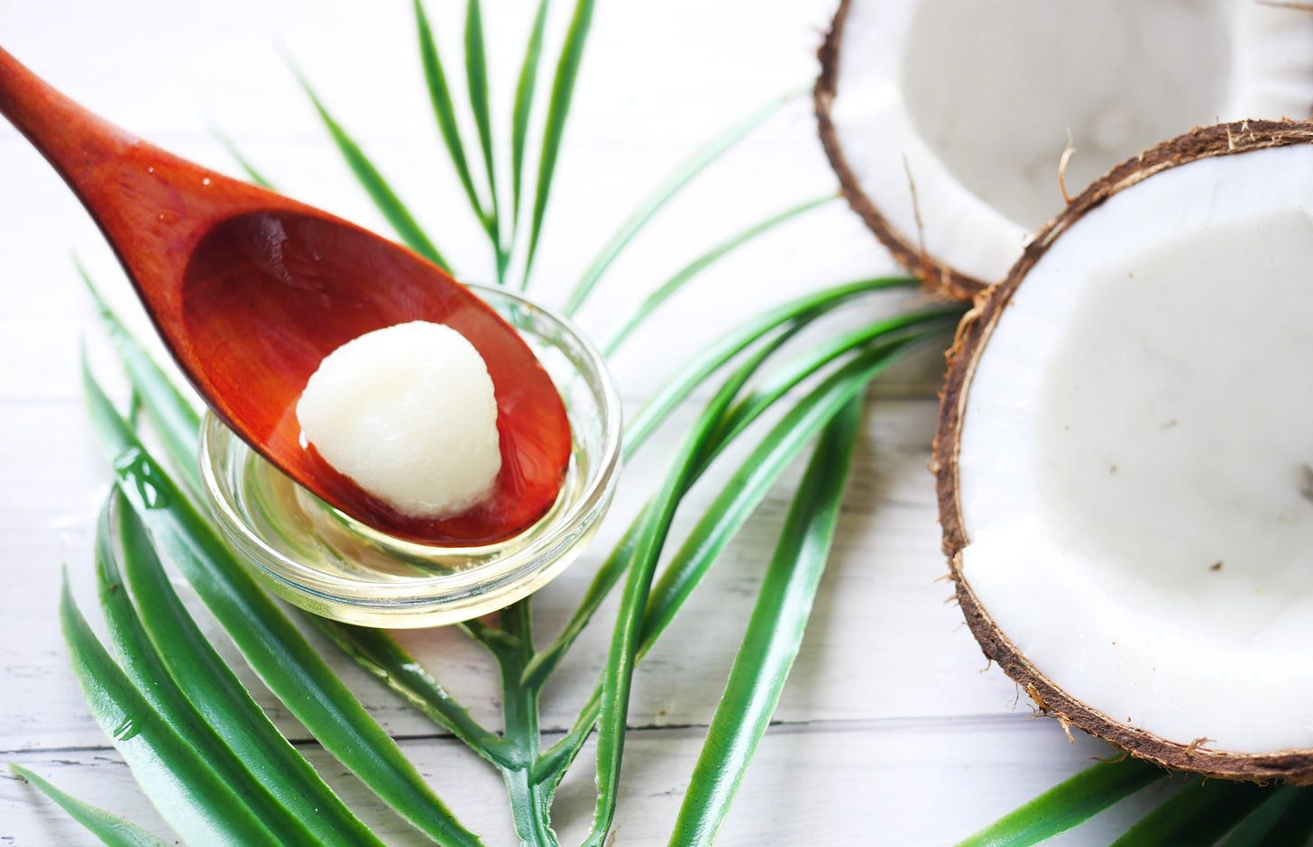 A halved coconut and a small bowl of coconut oil with a wooden spoon on a white surface, representing healthy saturated fats. Type Won Life™. A halved coconut and a small bowl of coconut oil with a wooden spoon on a white surface, representing healthy saturated fats. Type Won Life™.