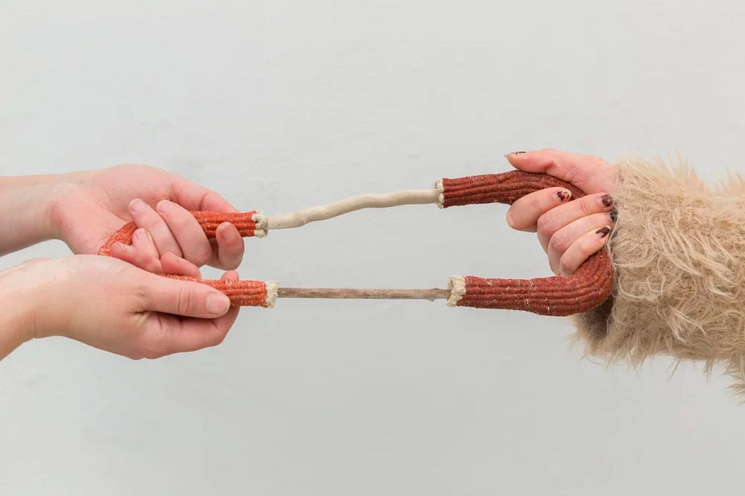 Two red, woven U-shaped basket forms connect two lengths of smooth, narrow drift wood, which are pressure-fit into the woven sockets. Ariana Martinez and Emma Berry tug at either end of the sculpture.