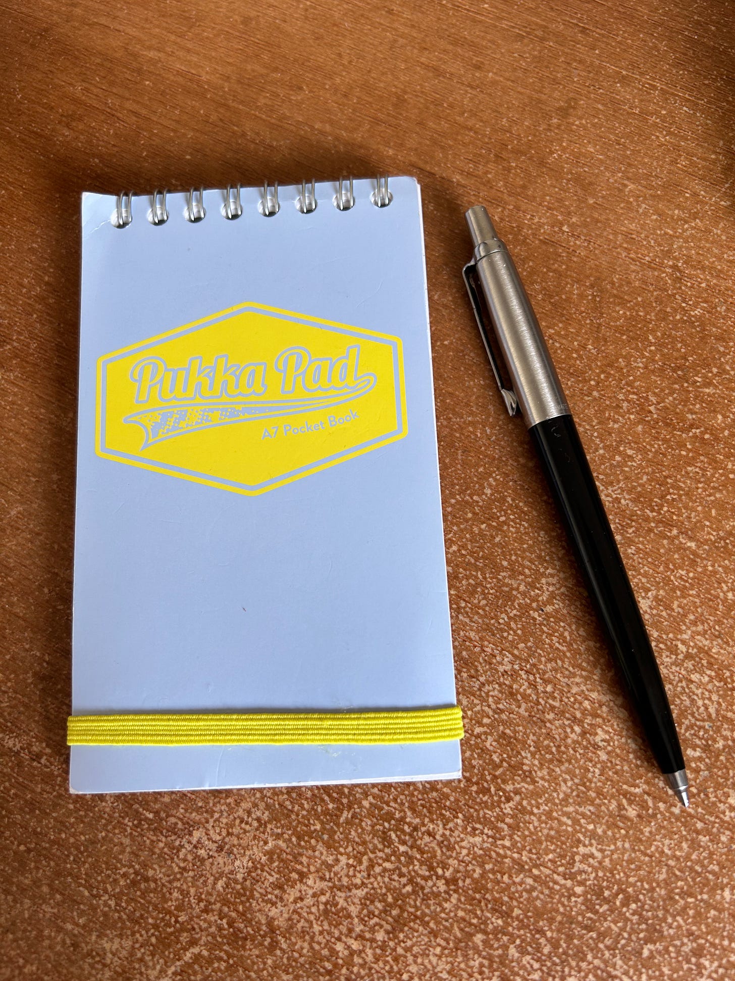 An A7 sized spiral bound notebook with a Parker Jotter pen alongside it lying on a desk.