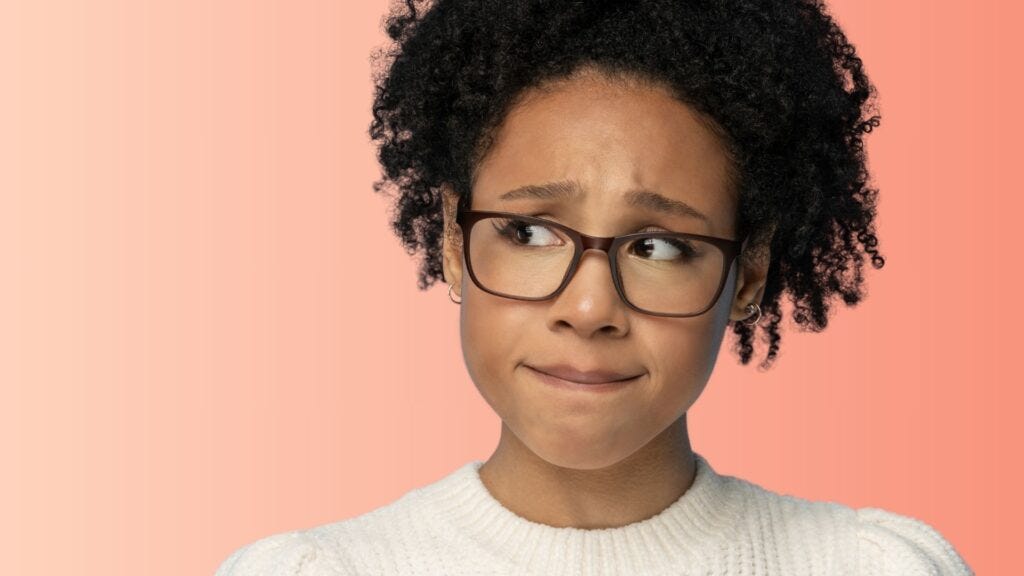 Young woman with curly hair and glasses looking uncertain and hesitant against a coral background, wearing a white sweater.