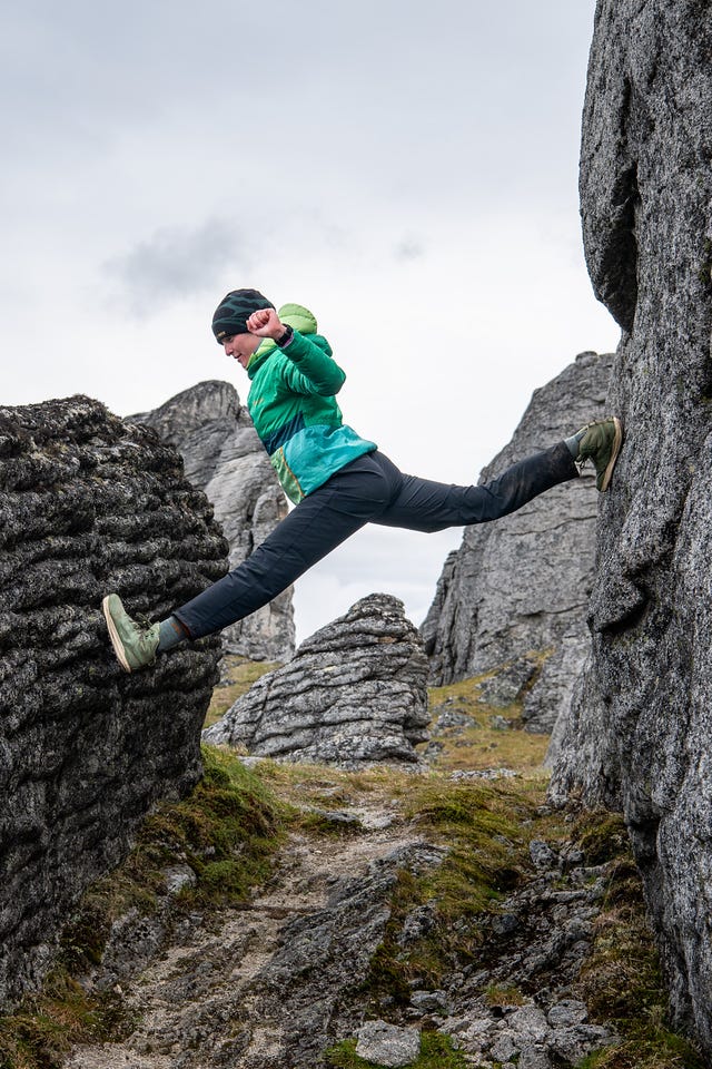 A hiker in a green jacket and black beanie braces between two granite walls, one foot high on each rock and legs stretched nearly to the splits, grinning, with more tors and overcast sky behind.