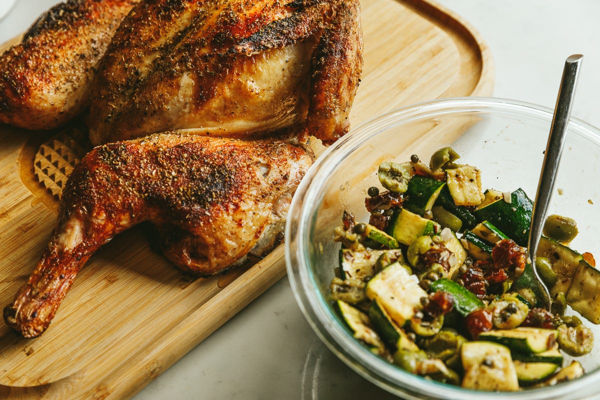 Grilled oregano spatchcock chicken on a cutting board with a warm zucchini salad.