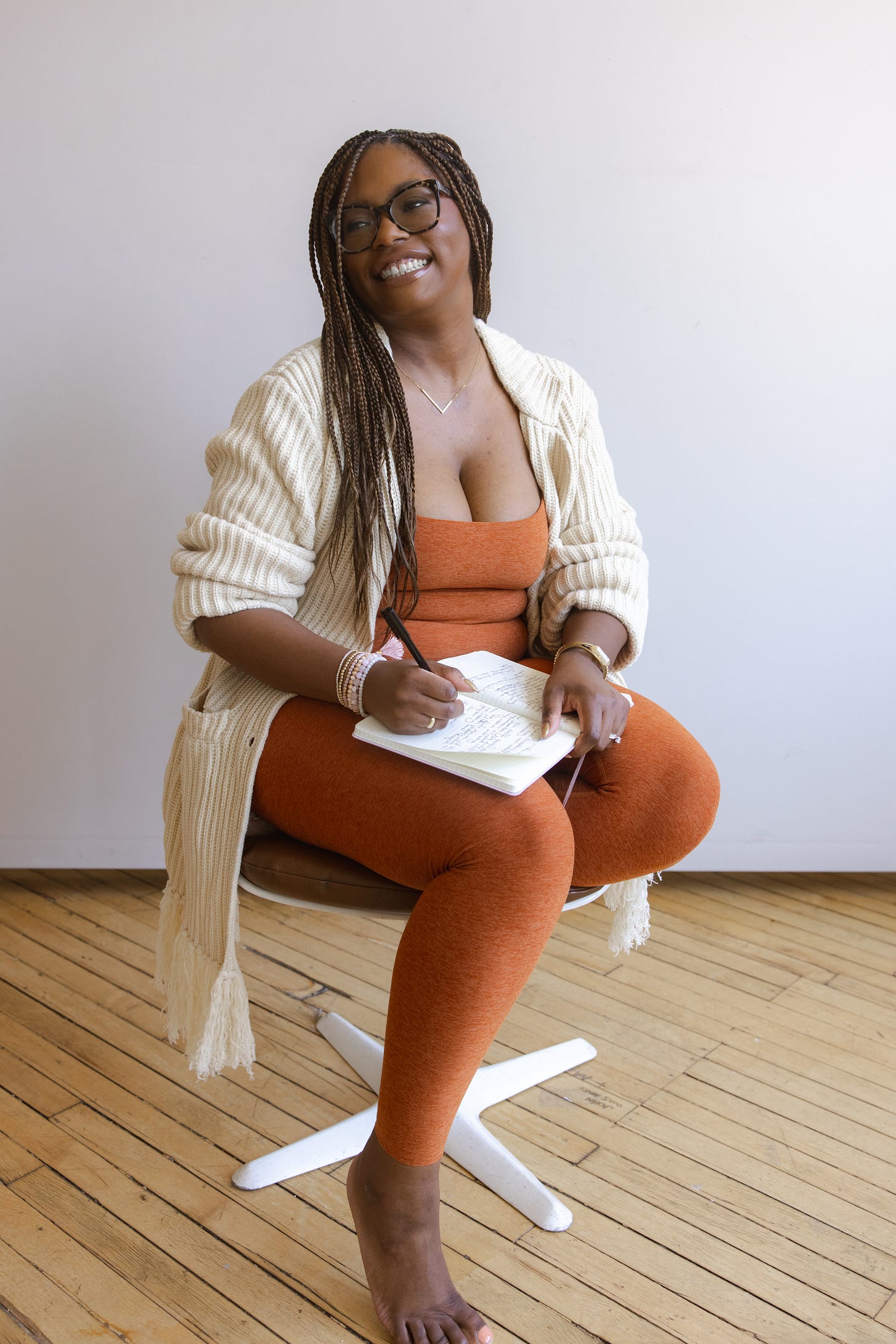 Picture of a Black woman wearing an orange tank top and matching leggings with a cream cardigan holding a journal in her lap, sitting and smiling off camera