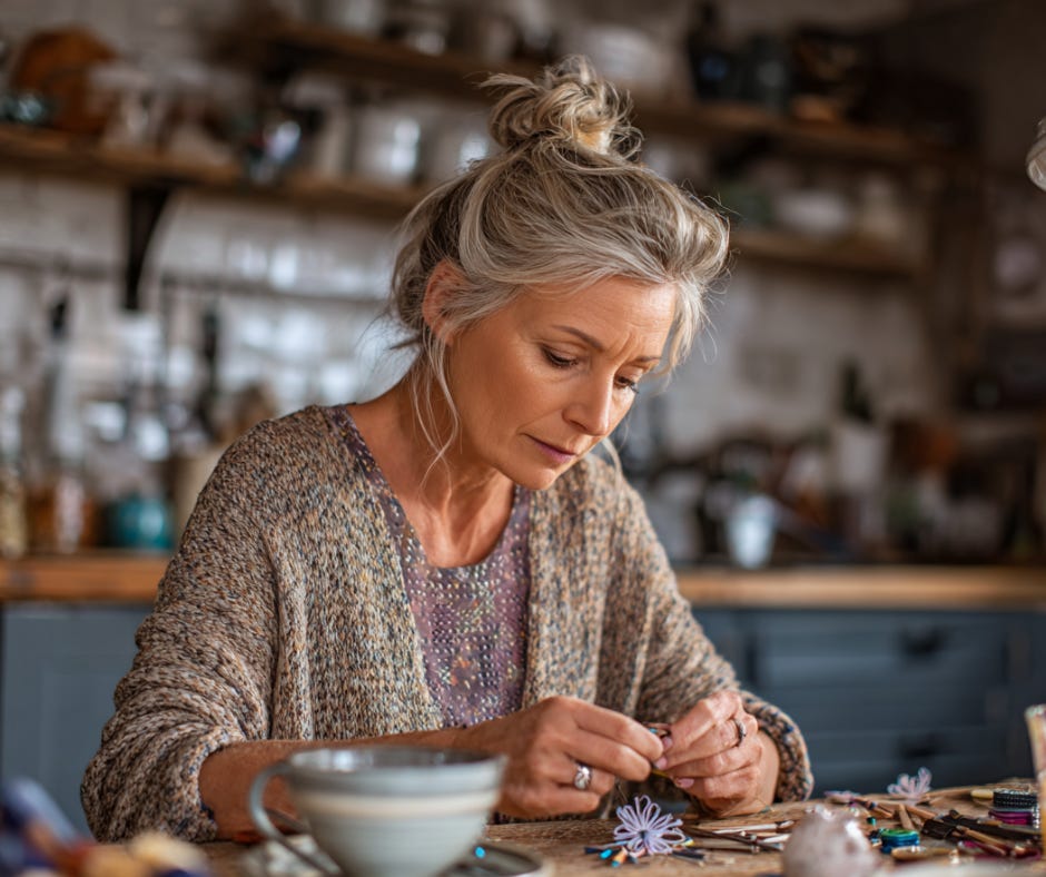 A woman in her 50s sits at a kitchen table deeply focused on crafting a handmade flower, surrounded by art supplies—capturing the quiet power of creative expression in midlife.