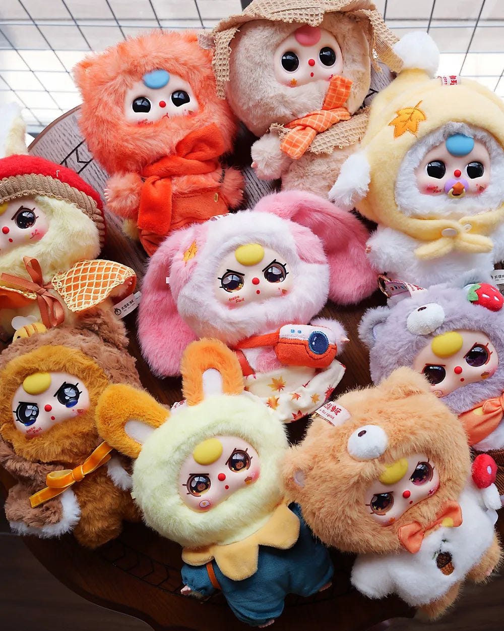 A tabletop arrangement of nine Baby Three “Autumn Fairy Tale” plush dolls, each wearing a different fluffy animal or seasonal outfit—fox, bear, bunny, lamb, and more—with signature big eyes, rosy cheeks, and bright accessories. 