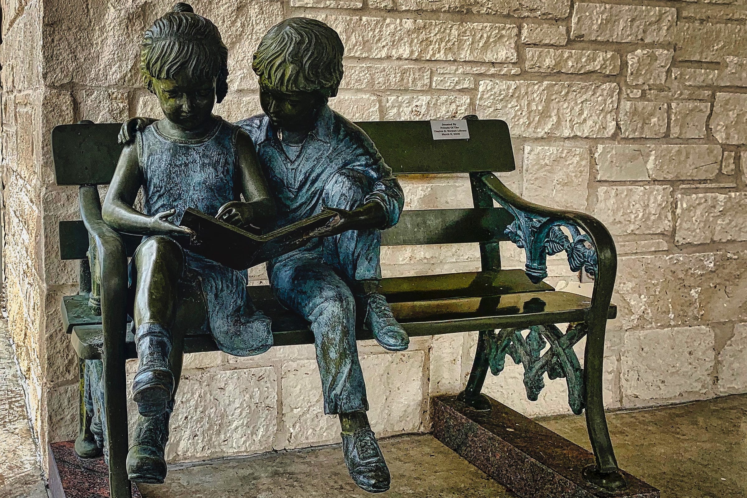 A sculpture of two young children on a park bench. The girl is holding a book while the boy looks on.