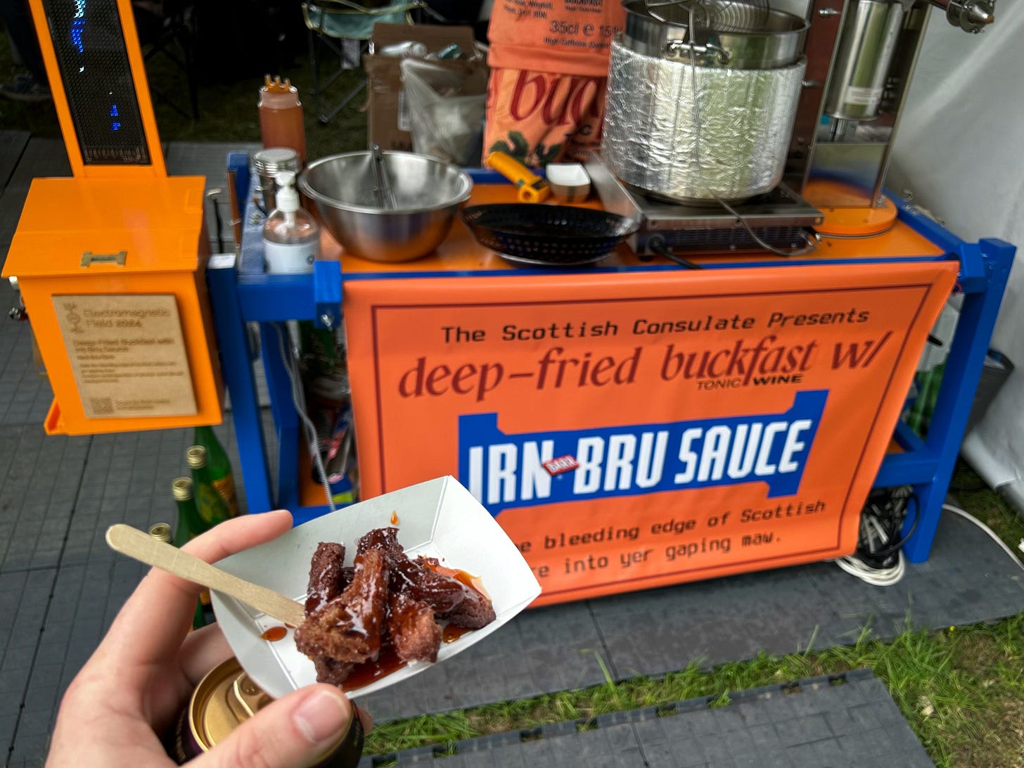 A photograph of a hand holding some deep fried Buckfast in front of a stand making it in EMF camp