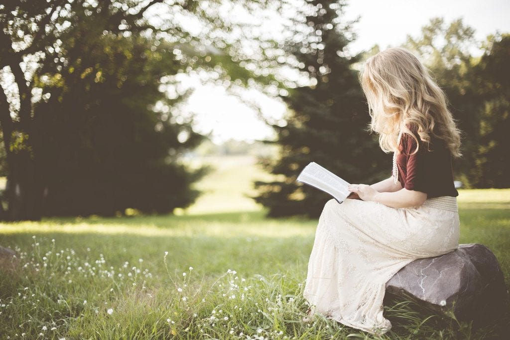 Long tousle-haired blonde woman in a long white skirt and burgundy 3/4 length sleeved sweater sitting on a rock reading fiction in the middle of a green grassy field with evergreen trees lining the horizon Long tousle-haired blonde woman in a long white skirt and burgundy 3/4 length sleeved sweater sitting on a rock reading fiction in the middle of a green grassy field with evergreen trees lining the horizon