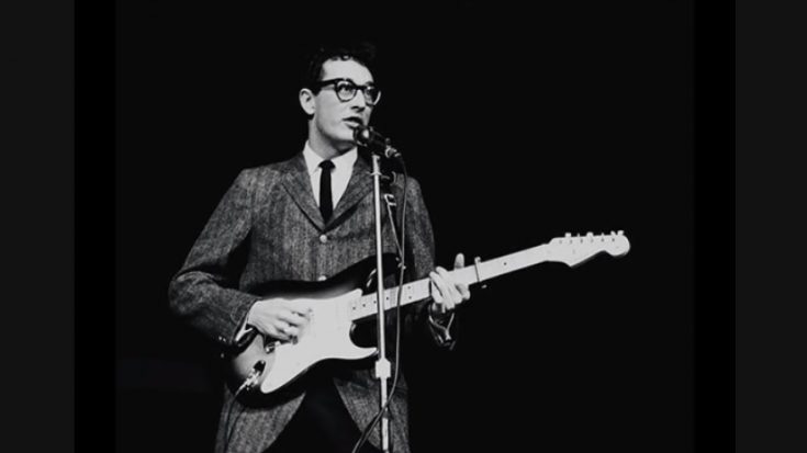 Buddy Holly, a white man holding an electric guitar and decked out in a suit and tie and some profoundly goofy glasses, preforming. It's a black and white picture and the background is completely black. 