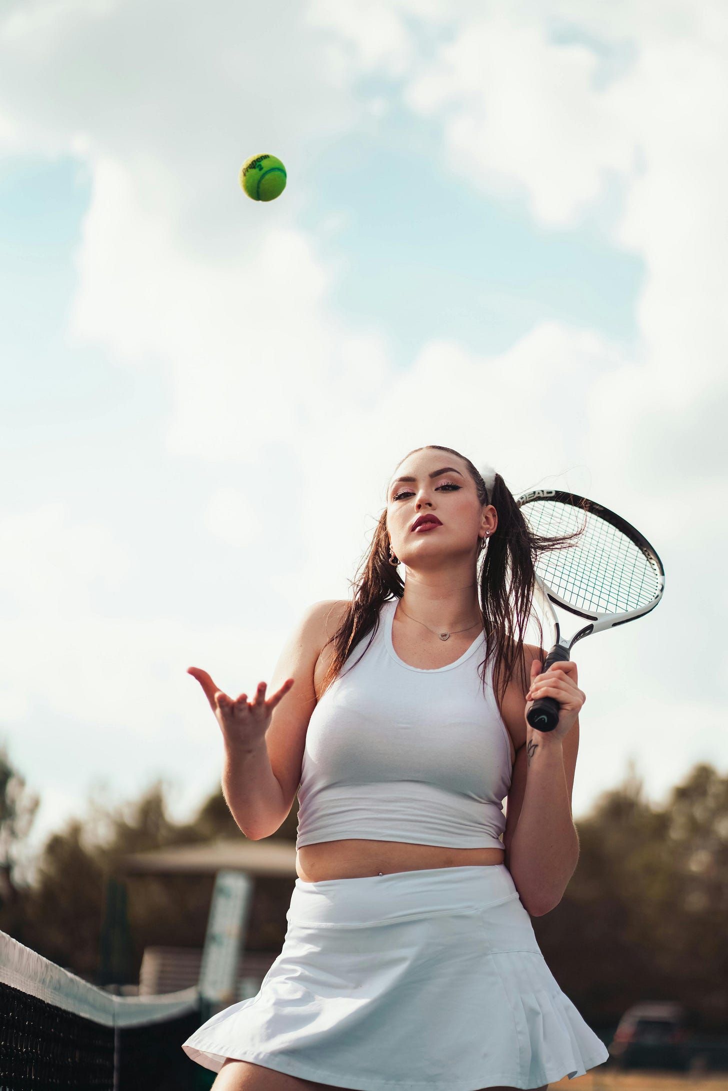 Young woman with dark hair wearing a white tennis skirt and crop top holding a tennis racquet over her left shoulder. She stares down at the camera. Behind her is a cloudy sky. Above her is a tennis ball which she has just thrown up in the air. Young woman with dark hair wearing a white tennis skirt and crop top holding a tennis racquet over her left shoulder. She stares down at the camera. Behind her is a cloudy sky. Above her is a tennis ball which she has just thrown up in the air.