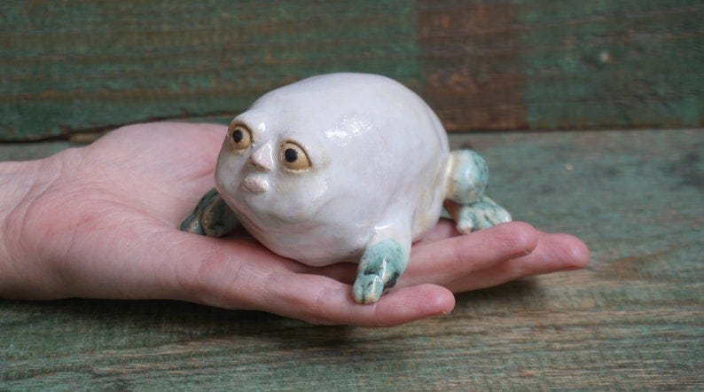 May include: A white ceramic frog figurine with green accents. The frog has a round body and large, expressive eyes. The frog is sitting on its hind legs with its front legs outstretched.