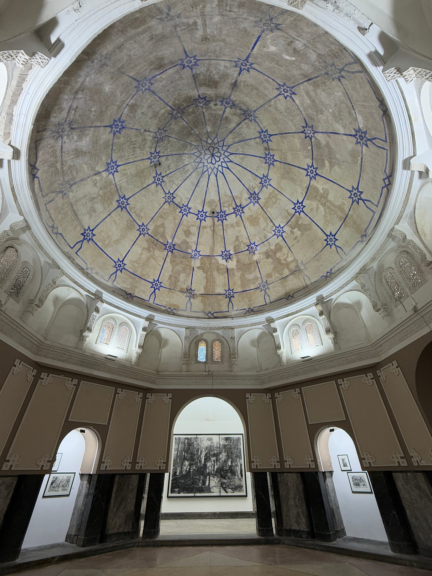 Interior view of Hammam El Bacha in Marrakech during the 1–54 art fair. A large circular dome with faded plaster and blue geometric star patterns dominates the ceiling. Below, arched openings frame exhibition walls with black-and-white artworks. The historic bathhouse architecture contrasts with the contemporary art installation. Interior view of Hammam El Bacha in Marrakech during the 1–54 art fair. A large circular dome with faded plaster and blue geometric star patterns dominates the ceiling. Below, arched openings frame exhibition walls with black-and-white artworks. The historic bathhouse architecture contrasts with the contemporary art installation.