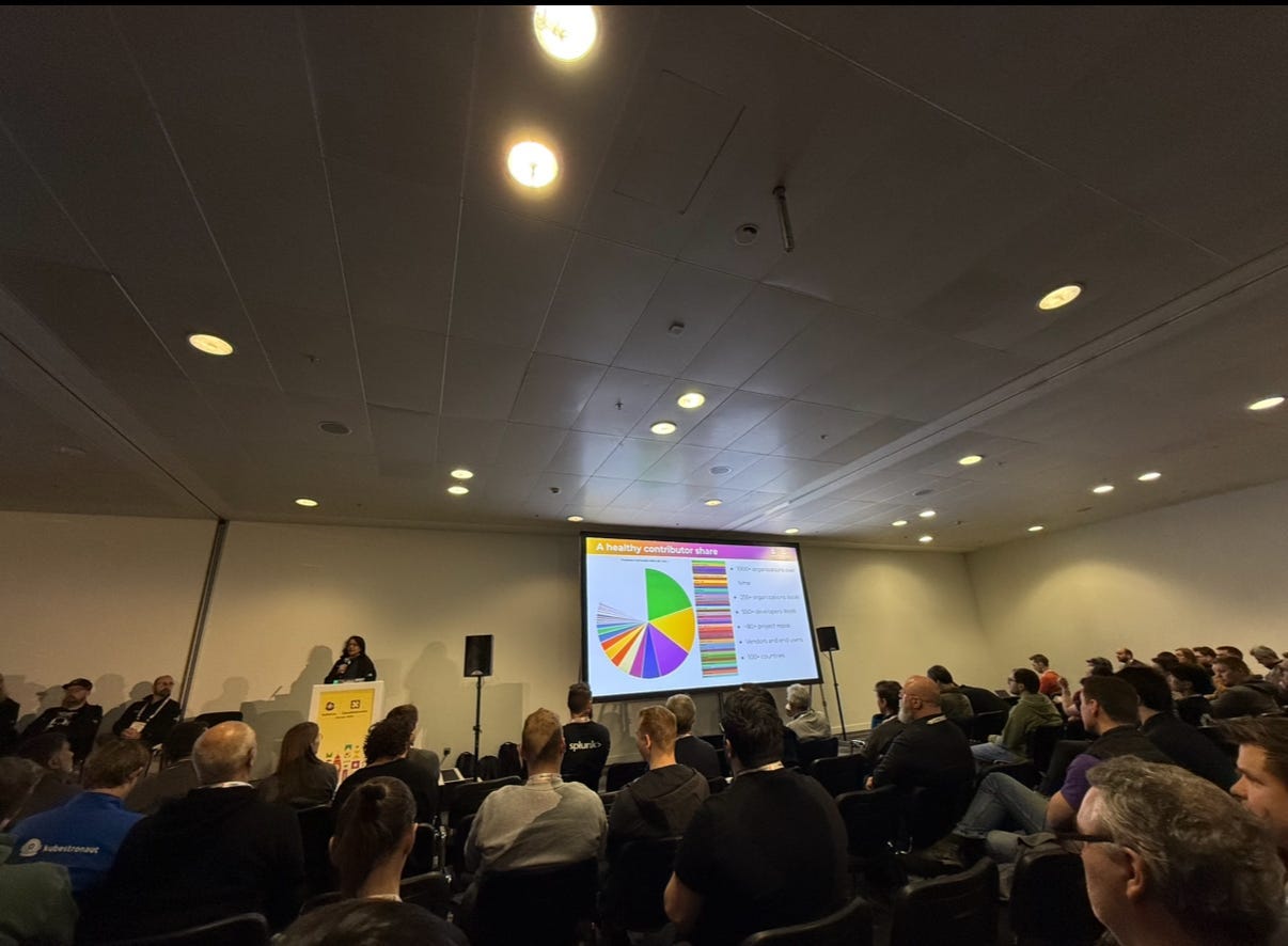 A conference room with attendees focused on a speaker at a podium. The speaker is presenting a slide titled “A healthy contributor share,” featuring a colorful pie chart and a list of categories such as organizations, designers hired, and project leads. The room is well-lit, with ceiling lights, and the audience is seated, facing the speaker and the presentation screen.