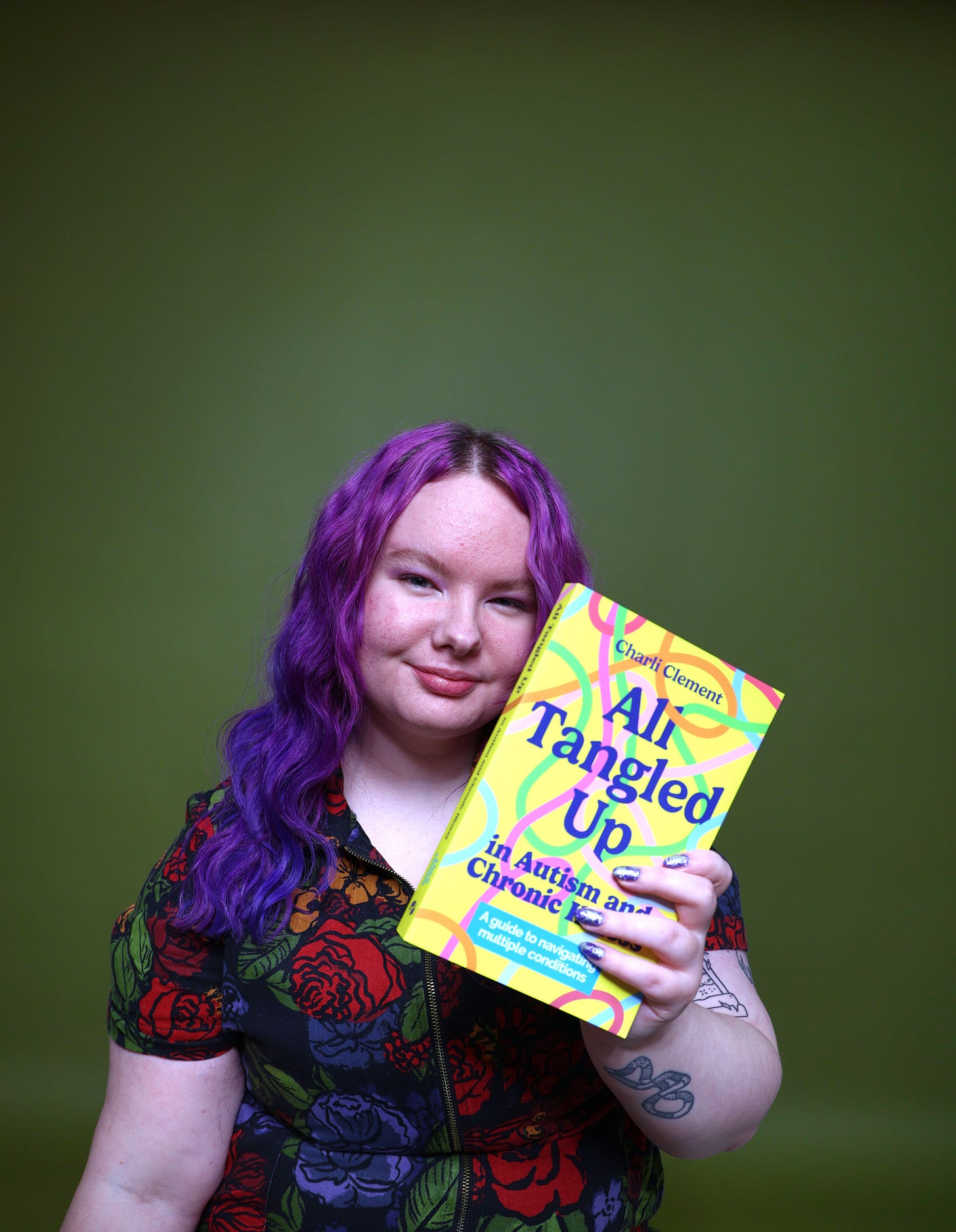 Charli is a white person with purple hair sat against an olive green background. They wear a floral jumpsuit and hold their book, All Tangled Up in Autism and Chronic Illness, which has a yellow cover with multicoloured squiggles 