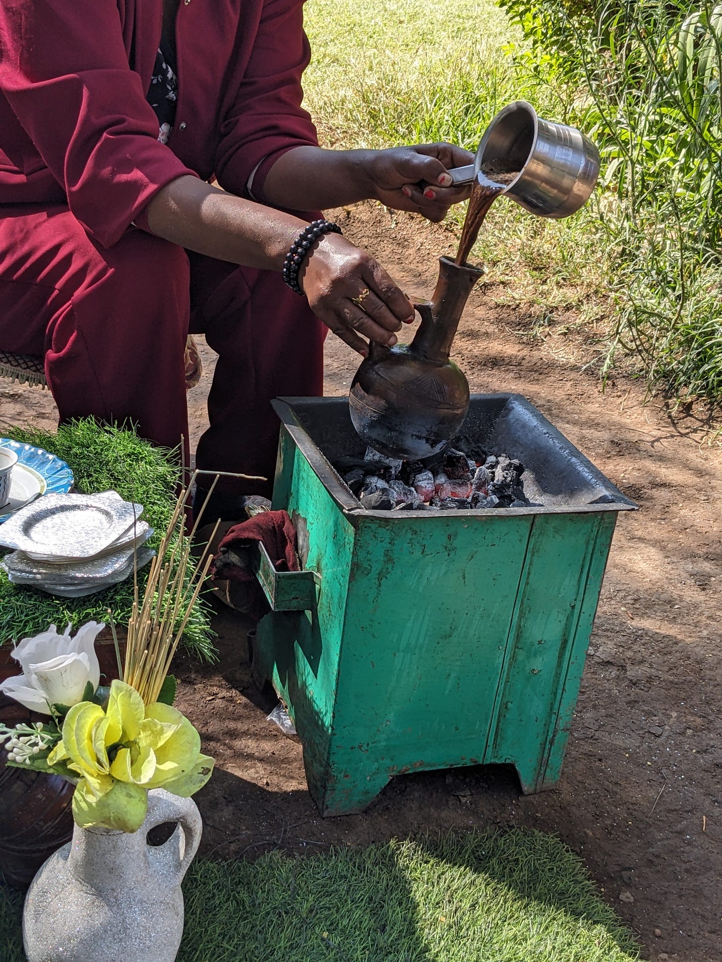 A black woman, wearing a red outfit, is pouring coffee over into a traditional clay pot. We don’t see her face. A black woman, wearing a red outfit, is pouring coffee over into a traditional clay pot. We don’t see her face.