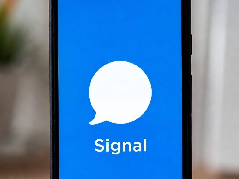 A photograph of a mobile phone displaying the Signal messaging app logo in a blurred indoor setting.