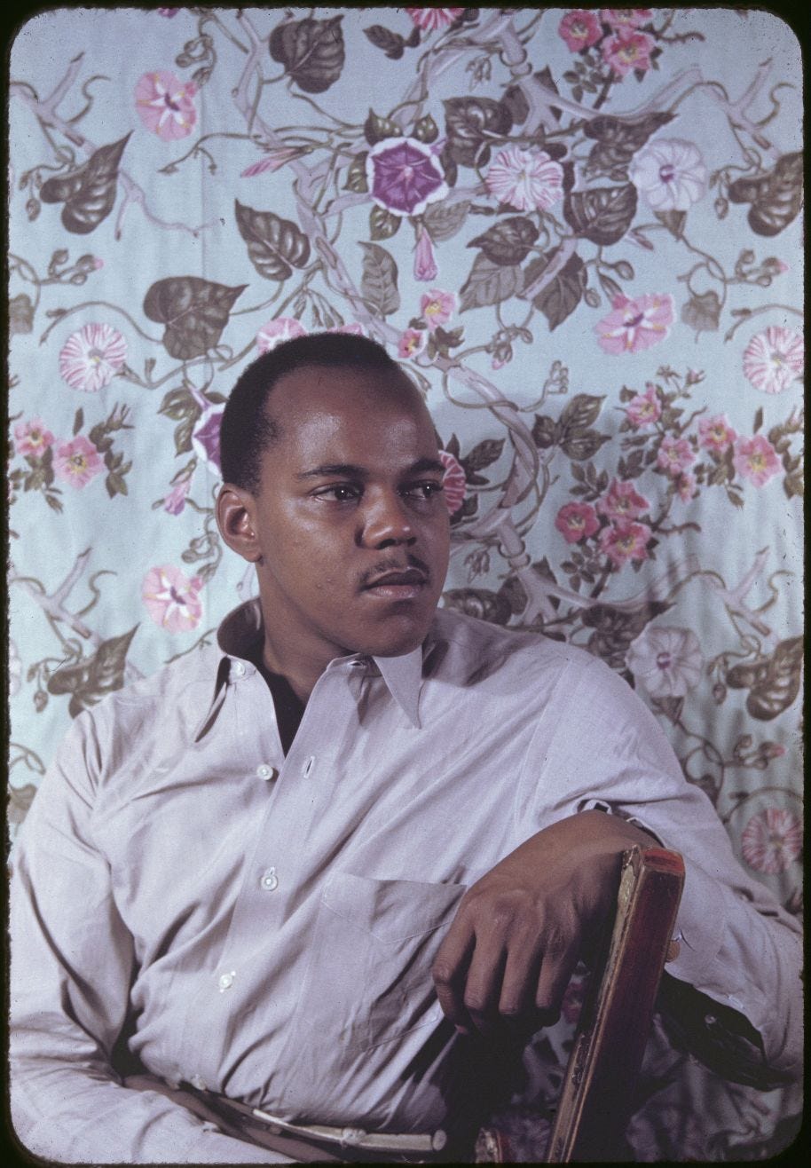 Portrait photograph of a young Black man wearing a light grey dress shirt, sitting in a wooden chair against a floral fabric backdrop