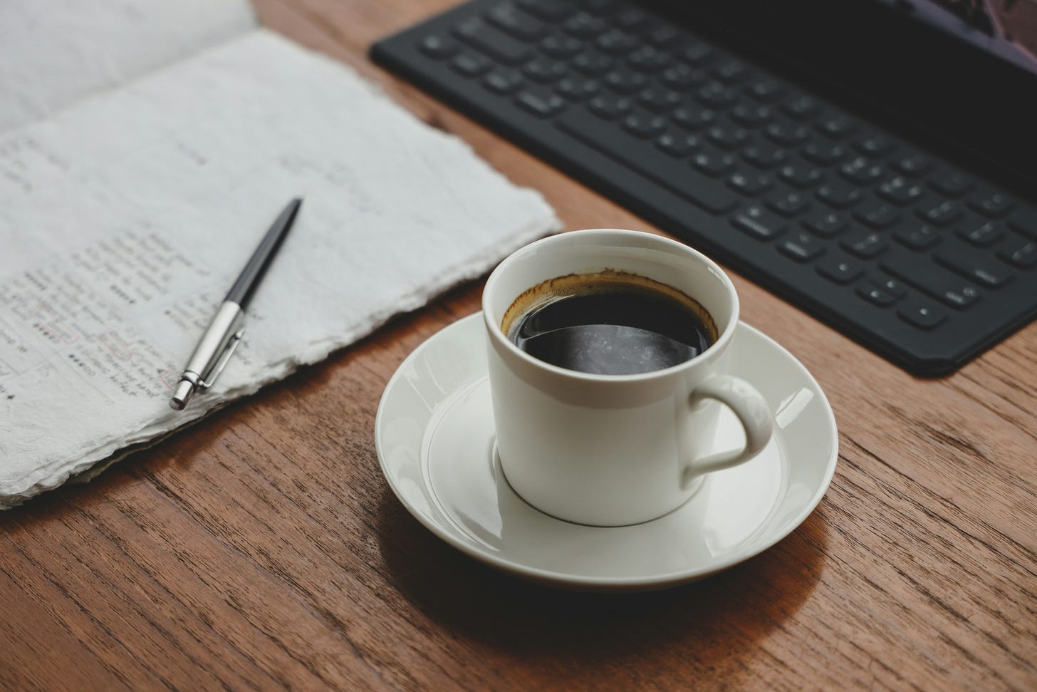 A white coffee cup on a saucer sits on a wooden desk alongside an open notebook and computer keyboard.