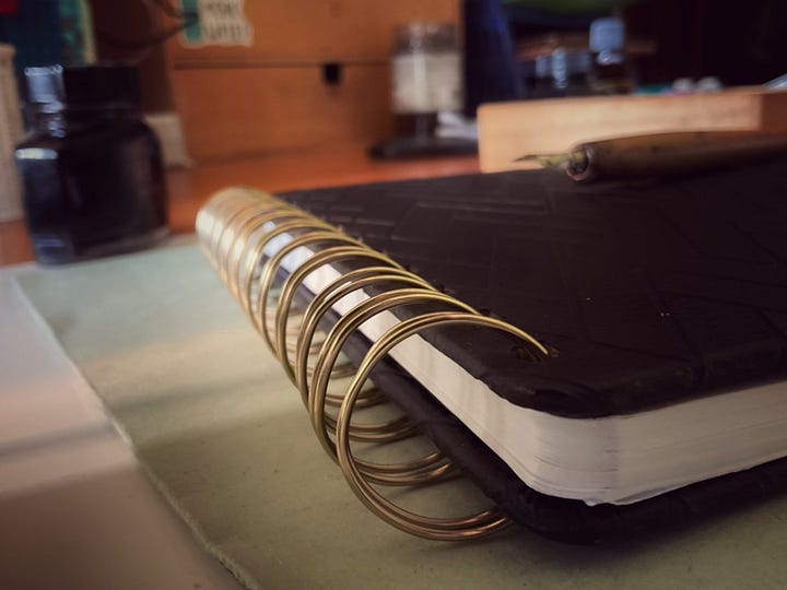 A split image displaying an artist's workspace. The left panel shows a black sketchbook with a herringbone texture, a wooden dip pen, and a Parker ink bottle sitting on a desk. The right panel is a close-up detail shot of the sketchbook's gold spiral binding, with the wooden pen slightly blurred in the background.