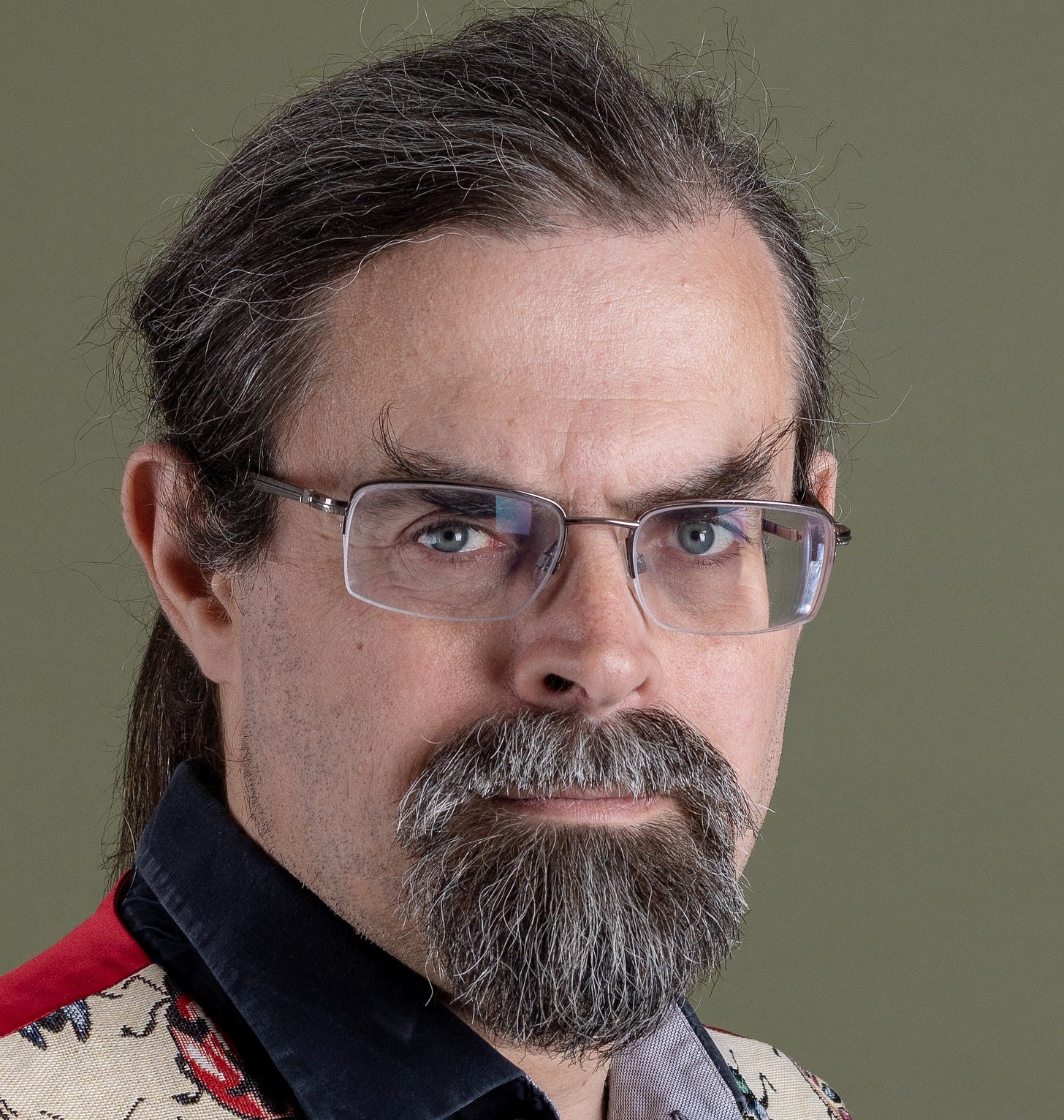 Studio portrait of a man with glasses, tied-back dark hair, and a grey beard, wearing a black shirt and a cream waistcoat patterned with colourful beetles.