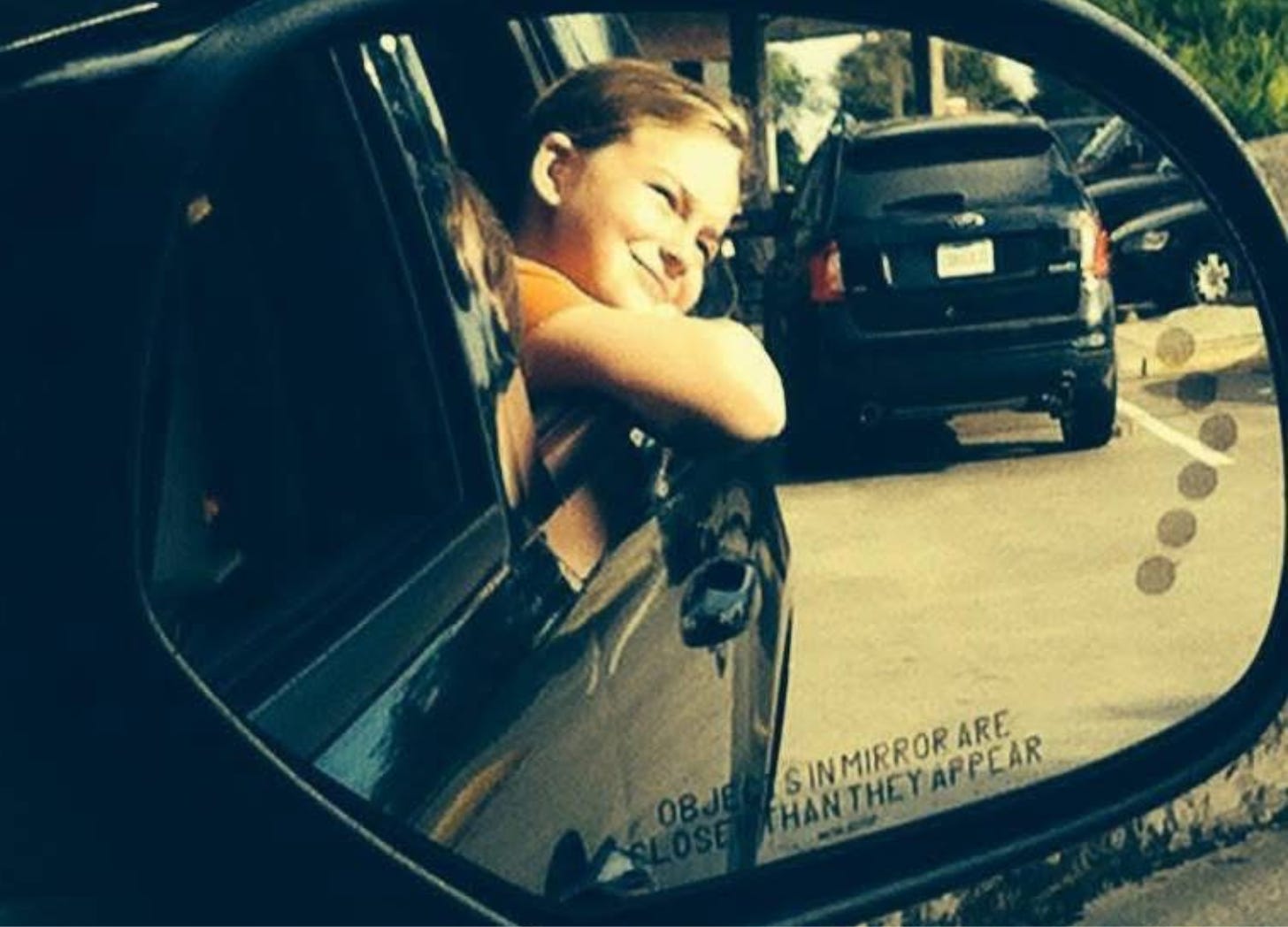 A child leaning out of a car window, captured in the reflection of a side mirror with the words "Objects in mirror are closer than they appear" clearly visible.