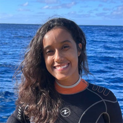 Mulher branca, com longos cabelos ondulados e castanhos, sorrindo e olhando para a câmera. Usa traje de mergulho preto, com detalhes claros e um cinto de mergulho e uma faixa turquesa no pulso. Fundo da imagem, oceano azul e céu claro com nuvens brancas esparsas.