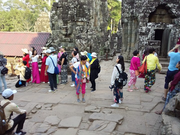 A side-by-side image. The left photo shows a large, crowded group of tourists in bright clothing standing on the ancient, uneven stone pathways of a Cambodian temple ruin. The right photo shows a busy dirt and paved road lined with tall trees, featuring large white tour buses, vans, and dozens of tourists walking around the drop-off zone.